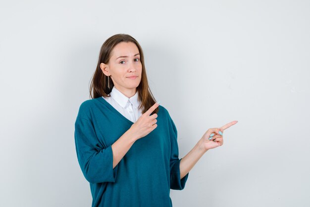 Porträt einer jungen Frau, die auf die obere rechte Ecke im Pullover über weißem Hemd zeigt und selbstbewusste Vorderansicht sieht