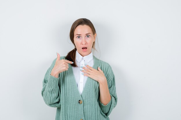 Porträt einer jungen Frau, die auf sich selbst zeigt, die Hand in Bluse, Strickjacke und überraschter Vorderansicht auf der Brust hält