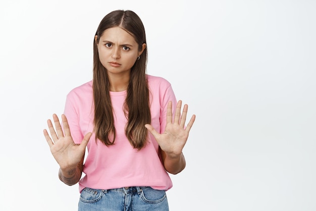 Porträt einer jungen Frau, die ernsthaft aussieht und die Hände nach vorne ausstreckt, zeigt Stopp-Ablehnungsgeste, die etwas mit besorgtem Gesicht auf weißem Hintergrund ablehnt