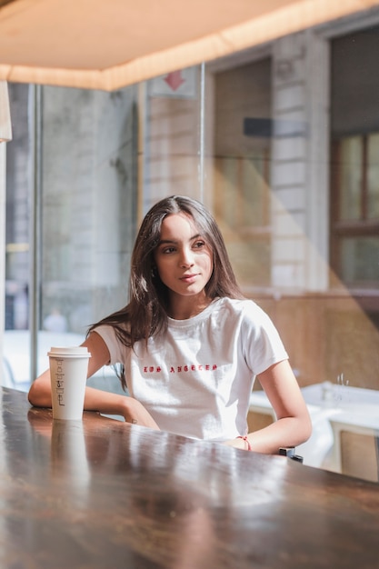 Porträt einer jungen Frau, die im Café mit Wegwerfkaffeetasse auf Tabelle sitzt