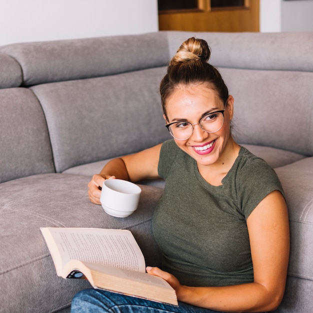 Porträt einer jungen Frau, die sich das Sofa hinsetzt, das Tasse Kaffee und Buch in den Händen hält