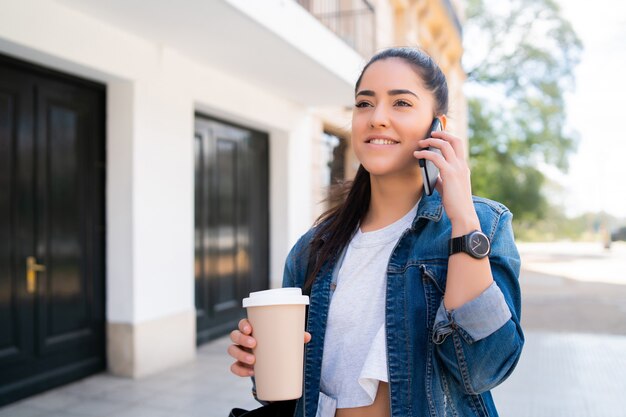 Porträt einer jungen Frau, die telefoniert und eine Tasse Kaffee hält, während sie draußen auf der Straße steht