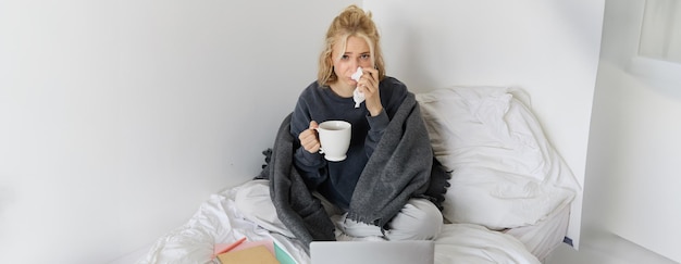 Porträt einer jungen Frau, die zu Hause sitzt, sich krank fühlt, Tee trinkt, Zeit im Bett verbringt