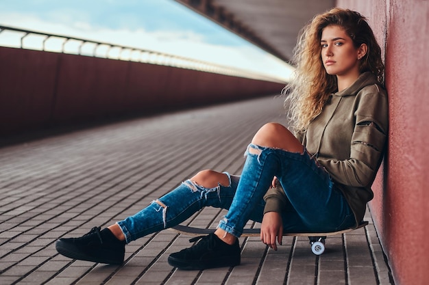 Porträt einer jungen Frau in einem Hoodie und zerrissenen Jeans, die sich an eine Wand lehnt, während sie auf einem Skateboard am Brückenfußweg sitzt.