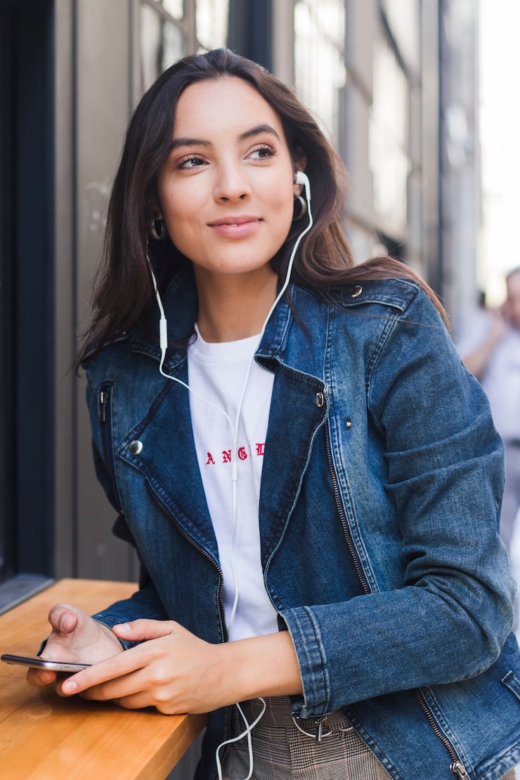 Kostenlose Foto porträt einer jungen frau in hörender musik der blauen denimjacke auf kopfhörer durch handy