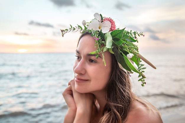 Porträt einer jungen Frau mit einer Zusammensetzung der Blumen auf ihrem Kopf vor dem Hintergrund des Ozeans bei Sonnenuntergang.