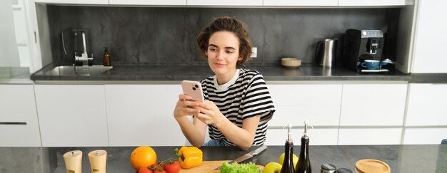 Kostenloses Foto porträt einer jungen frau mit smartphone, die in der küche sitzt und salat kocht und nach