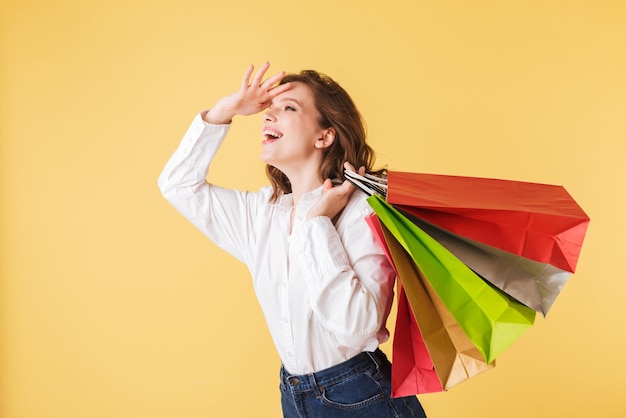 Kostenloses Foto porträt einer jungen, fröhlichen dame in hemd und jeansshorts, die mit bunten einkaufstüten in den händen steht und glücklich auf rosafarbenem hintergrund zur seite schaut