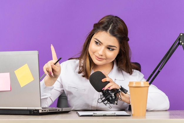 Porträt einer jungen Geschäftsfrau, die hinter dem Schreibtisch sitzt und mit dem Finger nach oben zeigt Foto in hoher Qualität