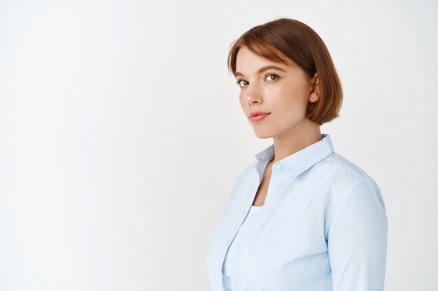 Porträt einer jungen Geschäftsfrau in Bluse mit kurzem Naturhaar, Blick und Lächeln, stehend gegen weiße Wand