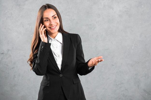 Porträt einer jungen glücklichen gestikulierenden Geschäftsfrau bei der Unterhaltung auf Mobiltelefon gegen grauen Hintergrund