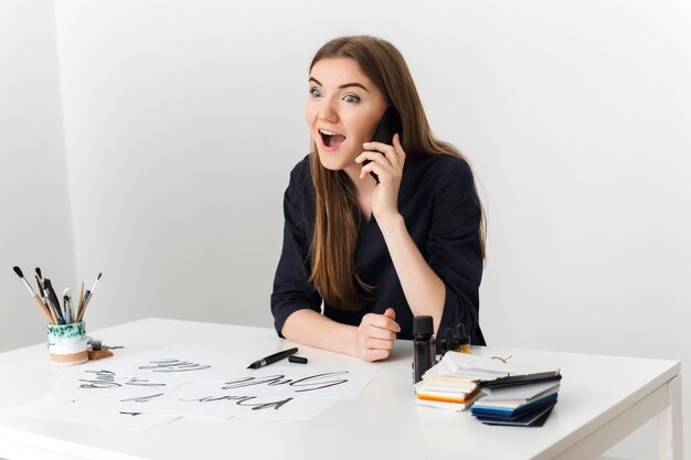 Porträt einer jungen hübschen Dame, die am weißen Schreibtisch sitzt und auf ihrem Handy spricht, während sie bewundernd isoliert beiseite schaut