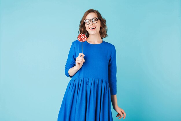 Porträt einer jungen lächelnden Dame mit Brille und Kleid, die mit Lollipop-Süßigkeiten in der Hand steht und glücklich in die Kamera blickt auf blauem Hintergrund