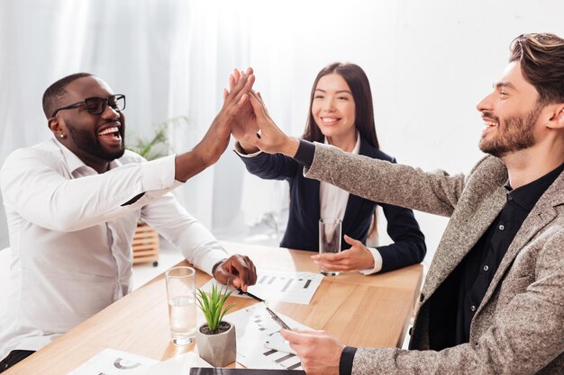 Porträt einer jungen multinationalen Gruppe von Geschäftspartnern, die am Tisch sitzen und sich freudig High Five geben, während sie isoliert im Büro zusammenarbeiten