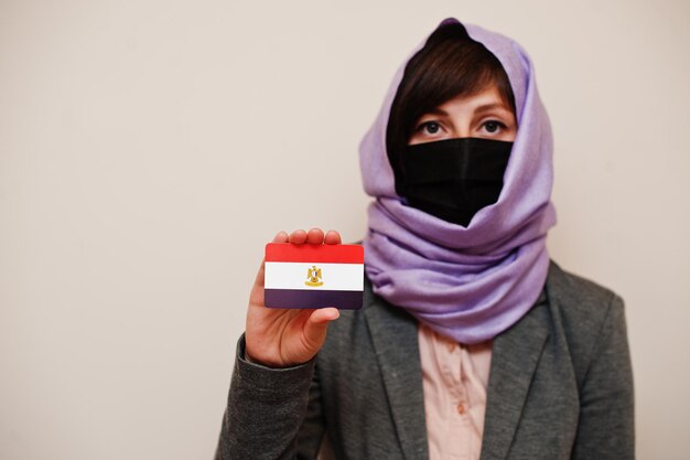 Porträt einer jungen muslimischen Frau, die formelle Kleidung trägt, schützt Gesichtsmaske und Hijab-Kopftuch, hält Ägypten-Flaggenkarte vor isoliertem Hintergrund Coronavirus-Landeskonzept