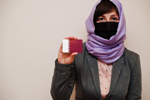 Porträt einer jungen muslimischen Frau mit formeller Kleidung schützt Gesichtsmaske und Hijab-Kopftuch und hält die Flaggenkarte von Katar vor isoliertem Hintergrund Coronavirus Country Concept