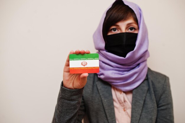Porträt einer jungen muslimischen Frau mit formeller Kleidung schützt Gesichtsmaske und Hijab-Kopftuch und hält die iranische Flaggenkarte vor isoliertem Hintergrund Coronavirus-Landeskonzept