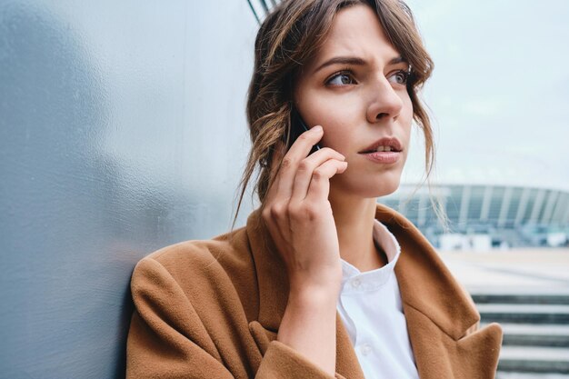 Porträt einer jungen, nachdenklichen Geschäftsfrau im Mantel, die auf dem Mobiltelefon im Freien spricht