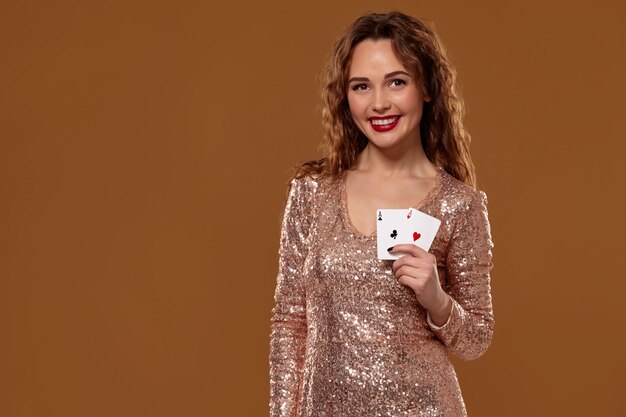Porträt einer jungen oder braunhaarigen Frau, die lächelt und ein Paar Asse hält, die ein goldenes Cocktailkleid auf braunem Hintergrund tragen. Casino-Konzept, Glücksspielindustrie