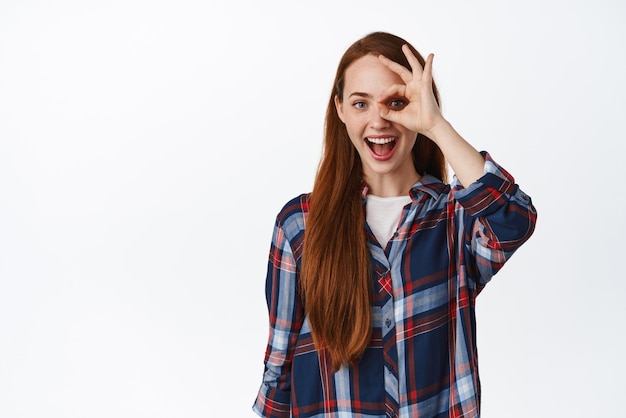 Porträt einer jungen rothaarigen Frau, die eine Okay-Zeichen-Null-Geste zeigt und ja sagt, sieht erstaunt und glücklich aus, wenn sie im karierten Hemd vor weißem Hintergrund steht