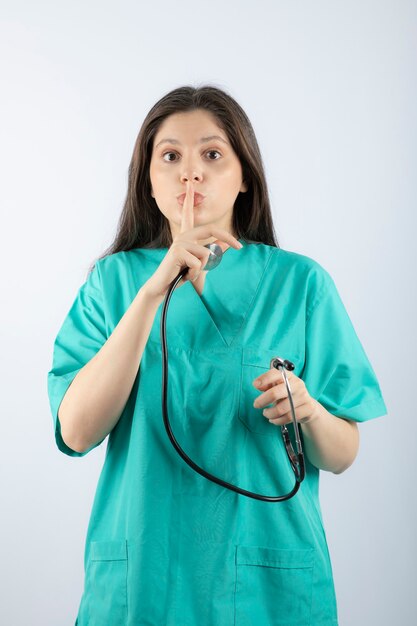 Porträt einer jungen Ärztin mit Stethoskop in Uniform, die ein stilles Zeichen zeigt.