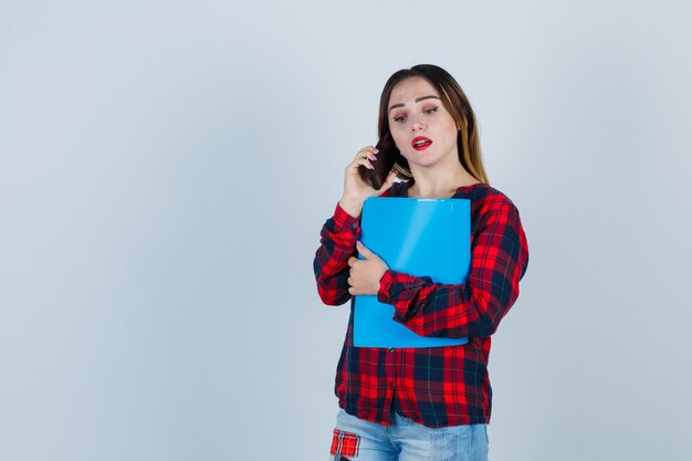 Porträt einer jungen schönen Frau, die einen Ordner hält, während sie in lässigem Hemd, Jeans und überraschter Vorderansicht telefoniert