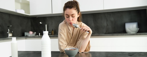 Kostenloses Foto porträt einer jungen schönen frau im bademantel, die müsli zum frühstück isst, lehnt sich an die küchenarbeitsplatte