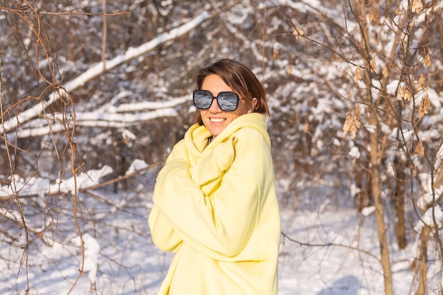 Porträt einer jungen schönen Frau in einem verschneiten Landschaftswinterwald an einem sonnigen Tag, gekleidet in einen gelben großen Pullover, mit Sonnenbrille, die Sonne und Schnee genießend