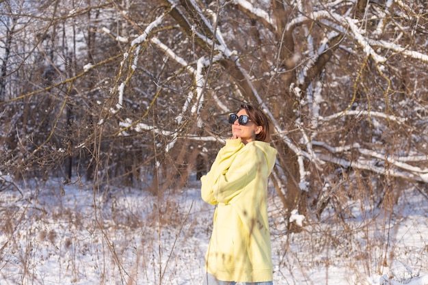 Porträt einer jungen schönen Frau in einem verschneiten Landschaftswinterwald an einem sonnigen Tag, gekleidet in einen gelben großen Pullover, mit Sonnenbrille, die Sonne und Schnee genießend