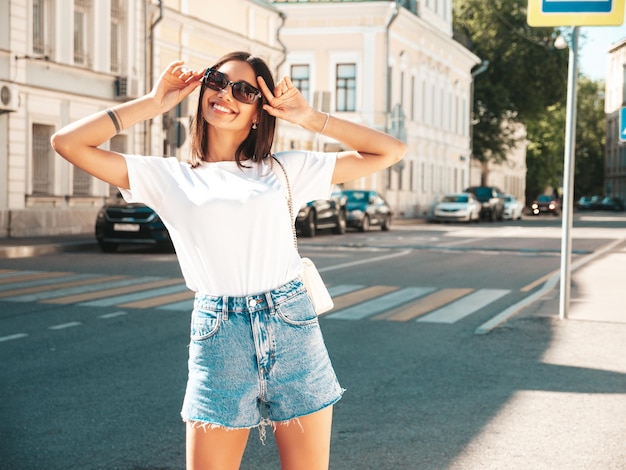 Porträt einer jungen, schönen, lächelnden Hipster-Frau in trendigen Sommer-Jeans-Shorts. Sexy sorgloses Modell, das bei Sonnenuntergang auf dem Straßenhintergrund posiert. Positives Modell im Freien mit Sonnenbrille