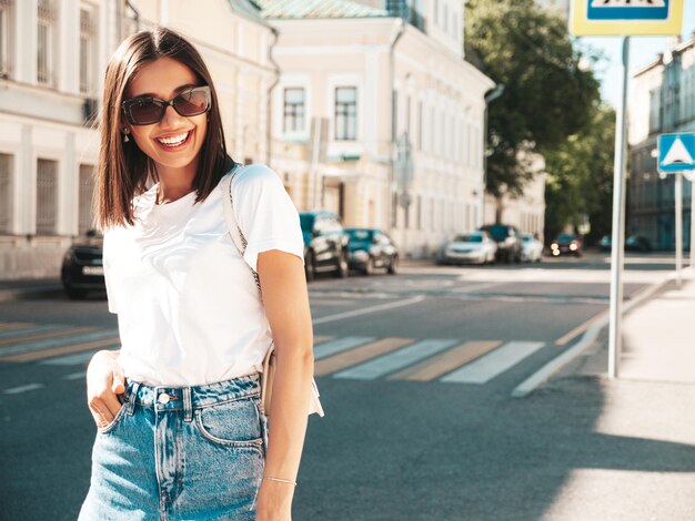 Porträt einer jungen, schönen, lächelnden Hipster-Frau in trendigen Sommer-Jeans-Shorts. Sexy sorgloses Modell, das bei Sonnenuntergang auf dem Straßenhintergrund posiert. Positives Modell im Freien mit Sonnenbrille