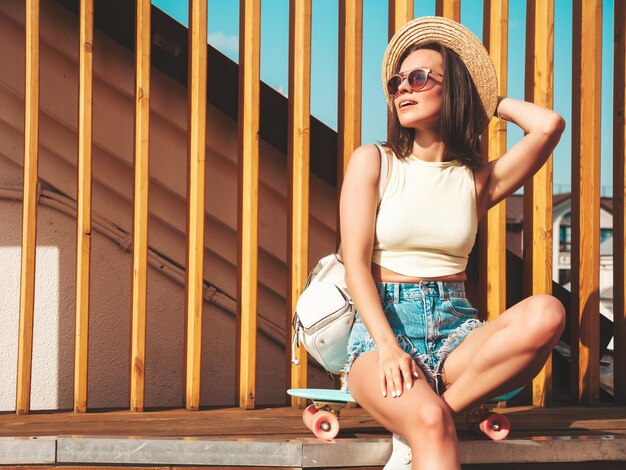 Porträt einer jungen, schönen, lächelnden Hipster-Frau in trendigen Sommerjeans, Shorts und Hut. Sexy unbeschwertes Modell, das bei Sonnenuntergang auf der Straße posiert. Positives Modell im Freien mit Aktentasche