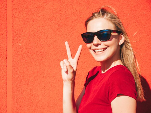Porträt einer jungen, schönen, lächelnden Hipster-Frau in trendiger Sommerkleidung Sexy sorglose Frau, die bei Sonnenuntergang in der Nähe einer roten Wand auf der Straße posiert Positives Modell im FreienZeigt Friedenszeichen