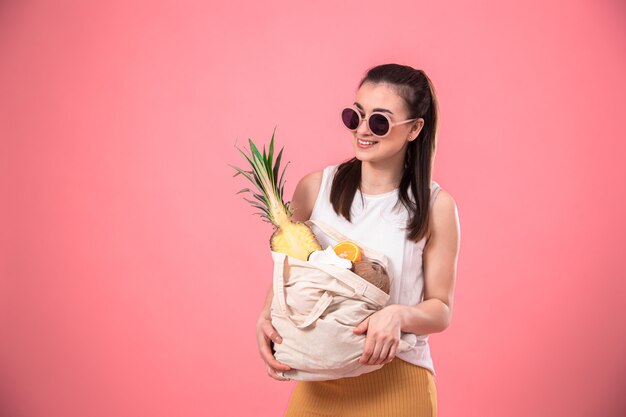 Porträt einer jungen stilvollen Frau in Sommerkleidung und Sonnenbrille, die eine ECO-Fruchttasche hält, auf rosa lokalisiert.