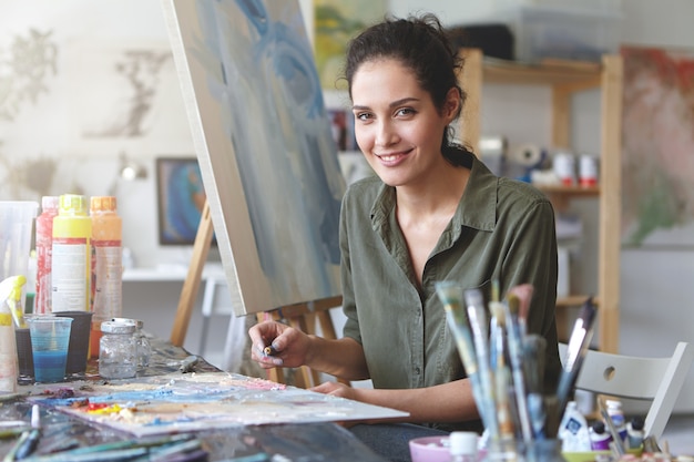Porträt einer jungen talentierten Künstlerin, die Skizzen mit hellen Ölen macht, auf Staffelei zeichnet, angenehmes Lächeln hat. Lächelnde Malerin, die mit ihrer Arbeit in der Werkstatt beschäftigt ist. Kunst, Kreativitätskonzept