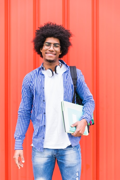 Porträt einer jungen Tragetasche des männlichen Studenten auf der Schulter und Büchern in der Hand, die gegen rote Wand stehen