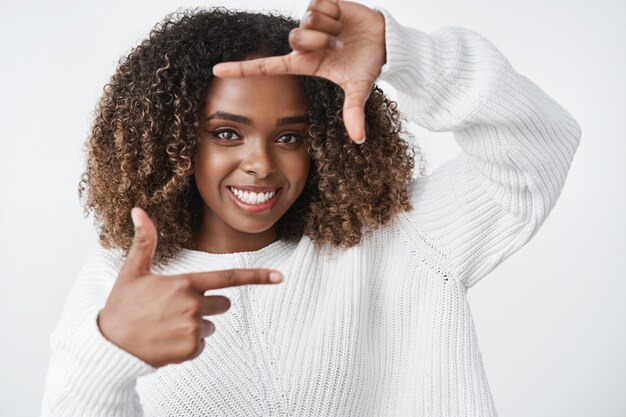 Porträt einer kreativen und enthusiastischen, gut aussehenden afrikanischen Frau mit lockigem Haar, die Rahmen mit den Händen zeigt und durch sie lächelnd und verträumt lächelt