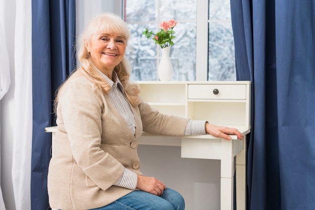 Porträt einer lächelnden älteren Frau, die vor Fenster nahe dem Schreibtisch sitzt