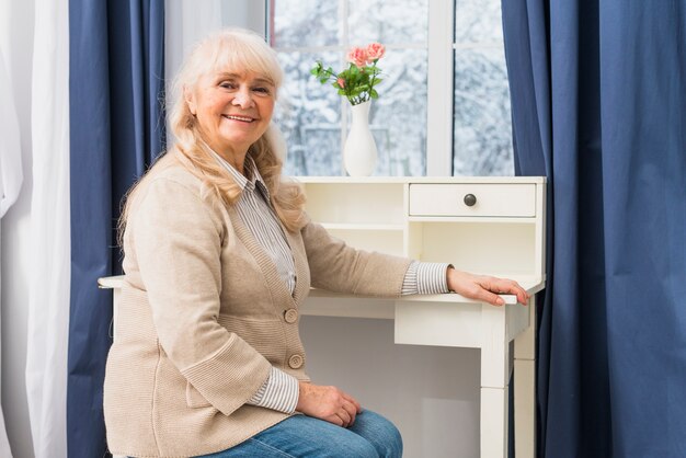 Porträt einer lächelnden älteren Frau, die vor Fenster nahe dem Schreibtisch sitzt