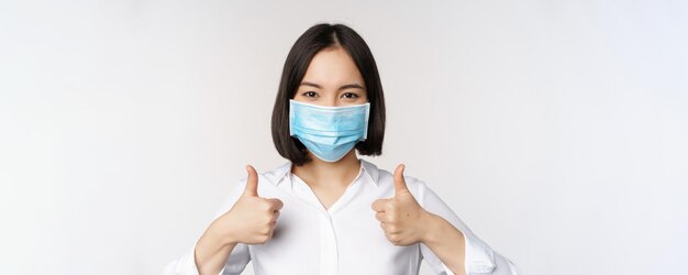 Porträt einer lächelnden asiatischen Bürodame in medizinischer Gesichtsmaske, die Daumen nach oben zeigt und etwas Stand empfiehlt