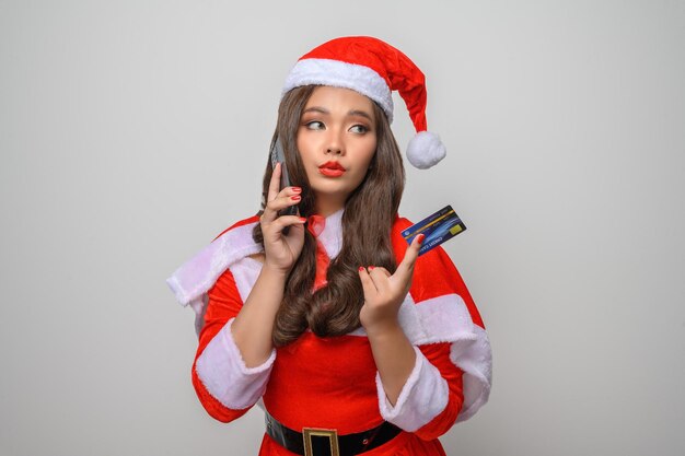 Porträt einer lächelnden asiatischen Frau im roten Weihnachtsmann-Kostüm, die Smartphone und Kreditkarte posiert