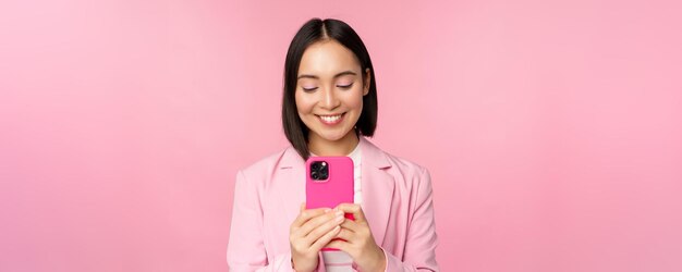 Porträt einer lächelnden asiatischen Unternehmensperson der Geschäftsfrau, die die Smartphone-Handyanwendung verwendet, die über rosa Hintergrund steht