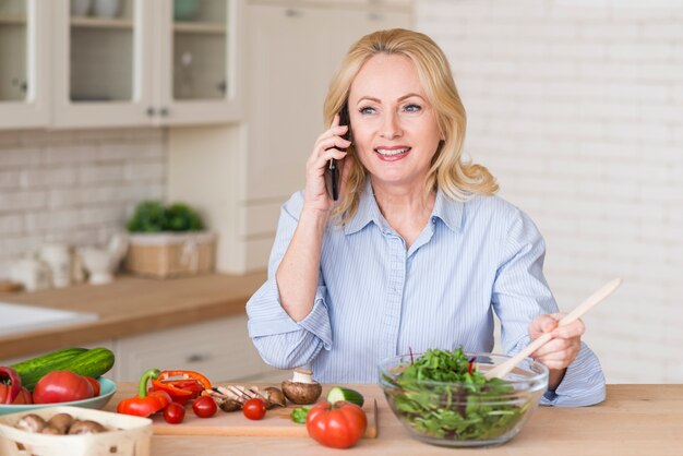 Porträt einer lächelnden blonden älteren Frau, die am Handy zubereitet den grünen Salat spricht