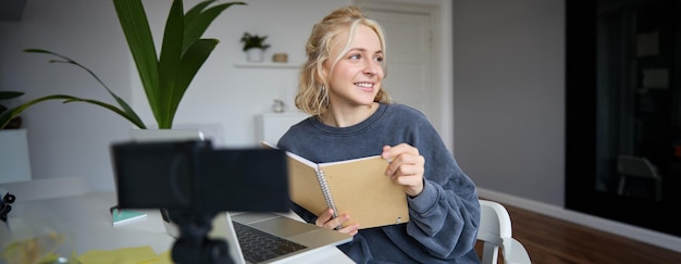 Kostenloses Foto porträt einer lächelnden blonden frau, die im schlafzimmer sitzt und einen laptop und eine digitalkamera verwendet, um videos aufzunehmen