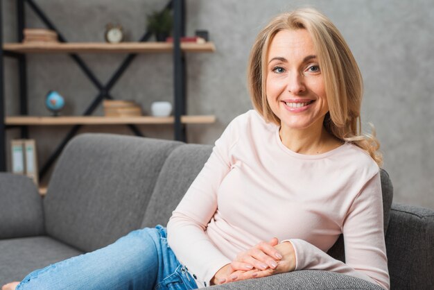 Porträt einer lächelnden blonden jungen Frau, die auf dem Sofa betrachtet Kamera sitzt