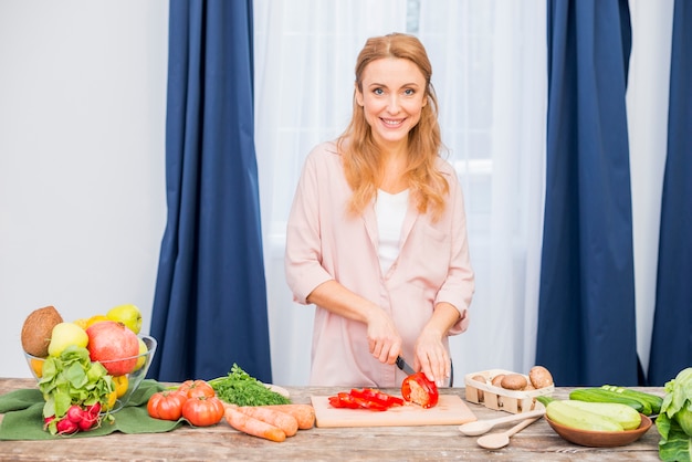 Kostenloses Foto porträt einer lächelnden blonden jungen frau, die den roten grünen pfeffer mit messer schneidet