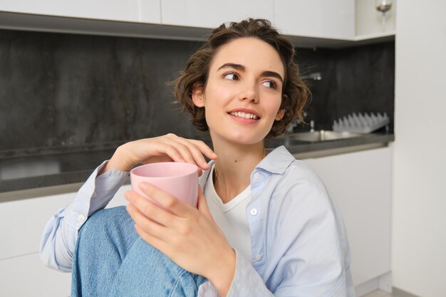 Porträt einer lächelnden brünetten Frau, die zu Hause sitzt, eine Tasse Tee in der Küche trinkt, entspannt und ihren freien Tag genießt