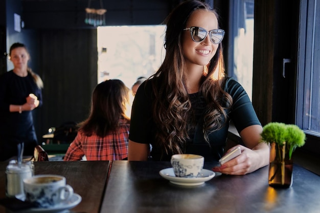 Porträt einer lächelnden brünetten Frau mit Sonnenbrille, trinkt Morgenkaffee in einem Café.