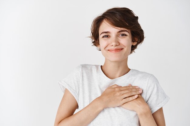 Porträt einer lächelnden europäischen Frau im T-Shirt, die das Herz berührt und lächelt, sich dankbar fühlt, Ihnen danke und gegen die weiße Wand steht standing