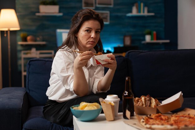 Porträt einer lächelnden Frau, die asiatische Nudeln zum Mitnehmen mit Essstäbchen auf dem Sofa im Wohnzimmer isst. Büroangestellter, der abends auf der Couch sitzt und chinesische Ramen-Boxen zum Mitnehmen und köstliches Junk Food genießt.